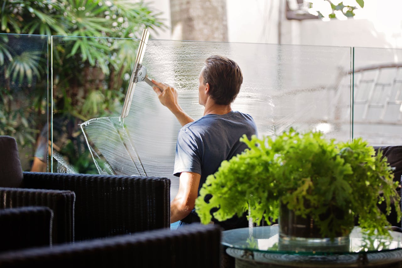 home-hero A man cleaning a glass barrier outdoors with a squeegee, surrounded by greenery.
